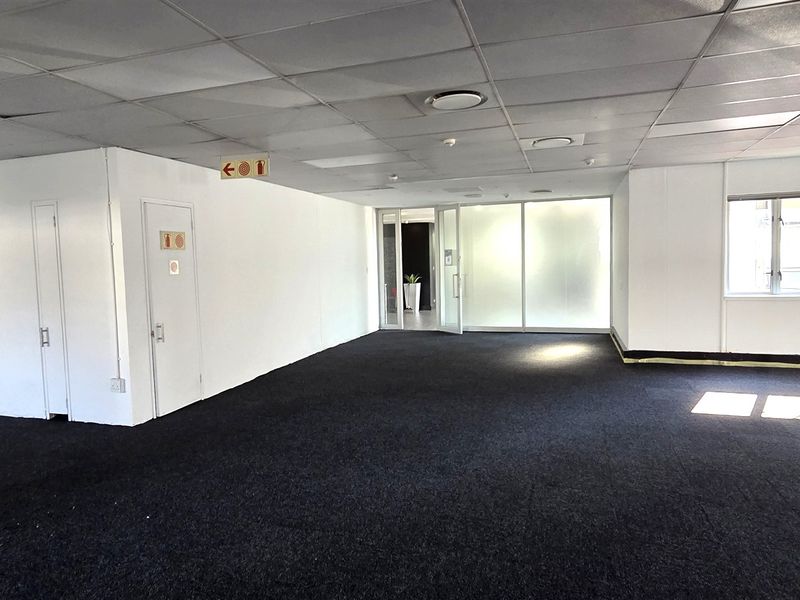 An open-plan office space featuring a suspended ceiling, recessed lighting, and dark carpeting, with glass partitioning and natural light from side windows.