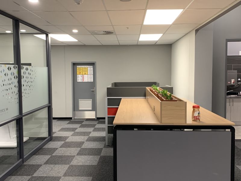 Modern industrial office space featuring glass partitions, carpeted flooring, suspended ceiling with recessed lighting, and a built-in workstation area.