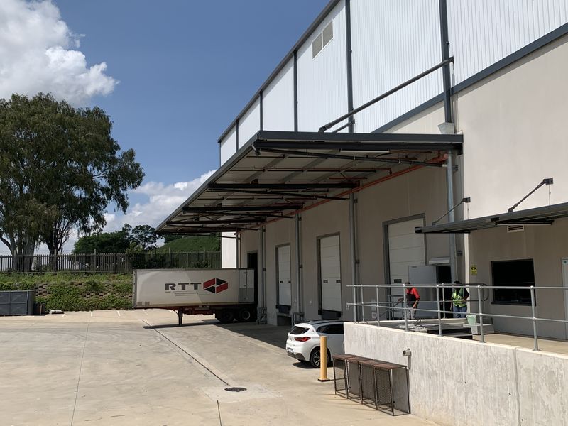 Industrial warehouse exterior featuring multiple loading docks with roller shutter doors, a concrete yard, and a protective metal canopy.