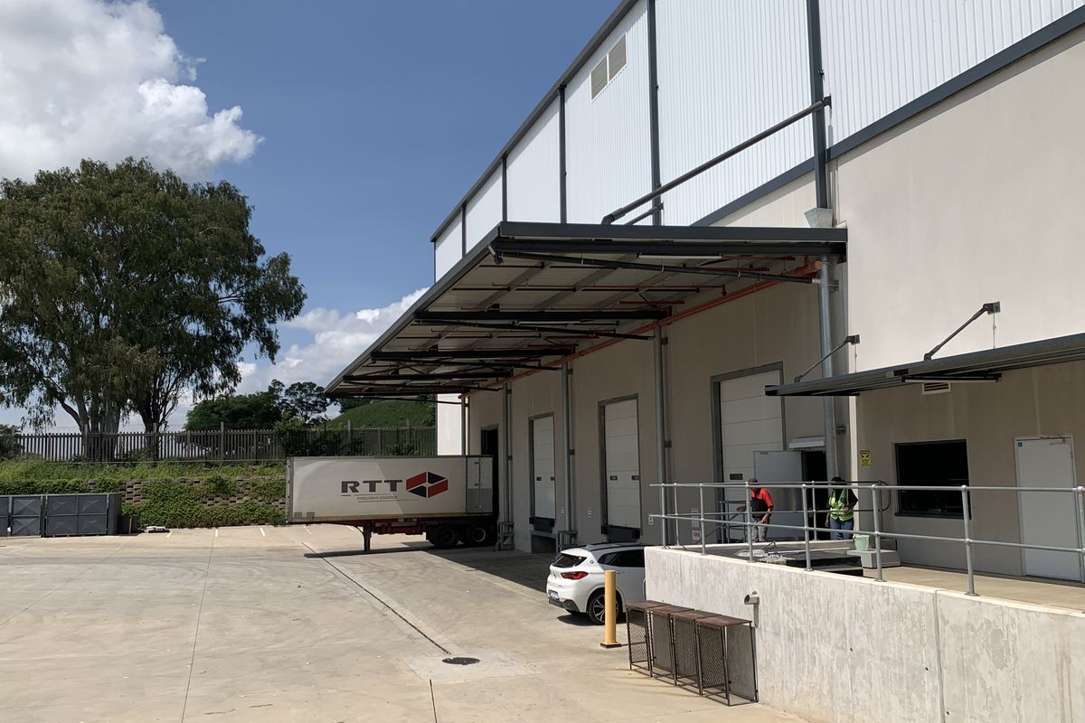 Industrial warehouse exterior featuring multiple loading docks with roller shutter doors, a concrete yard, and a protective metal canopy.