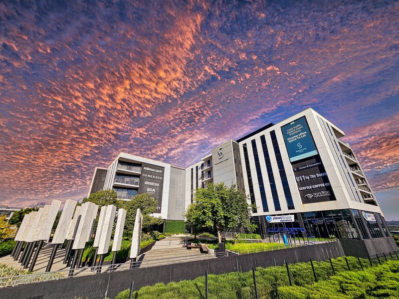 A modern multi-story office building featuring a contemporary glass facade, landscaped courtyard, and prominent commercial signage under a dramatic sunset sky.