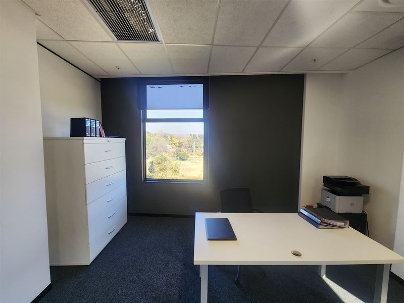 A functional office space featuring a suspended ceiling with integrated air conditioning, carpeted flooring, and a large window providing natural light and exterior views.