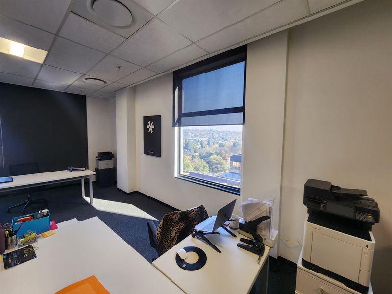A professional office space featuring a suspended ceiling, carpeted flooring, and a large window with roller blinds offering an elevated view.
