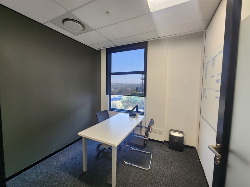 A professional office space featuring carpeted flooring, a suspended ceiling with recessed lighting, a built-in whiteboard, and a window with a roller blind.