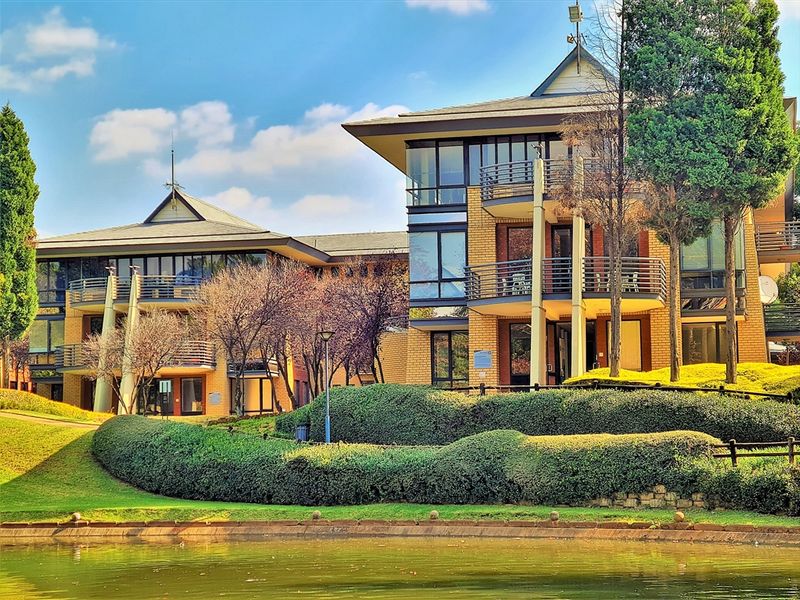 A well-maintained office complex featuring brick facades, multiple balconies with metal railings, and landscaped grounds overlooking a water feature.