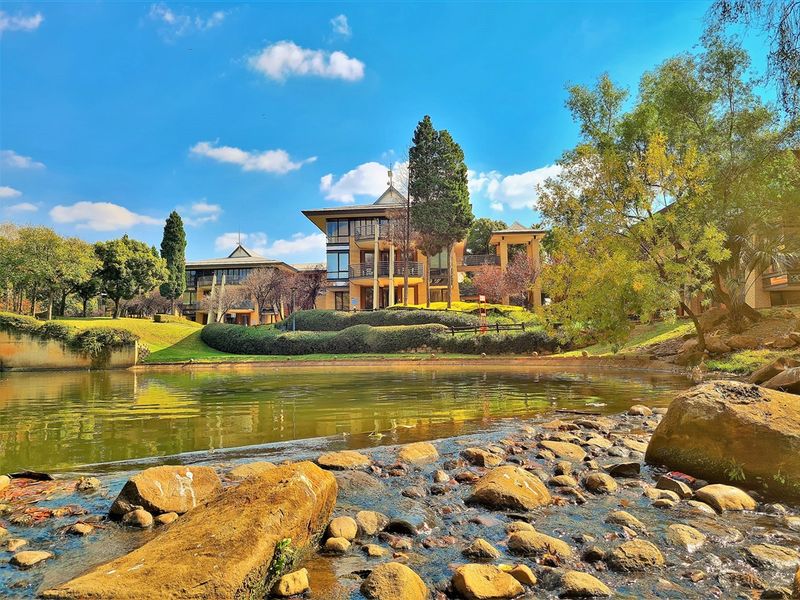 A scenic view of the office complex featuring multi-level architecture, balconies, and well-maintained landscaped gardens overlooking a tranquil pond and rocky stream.