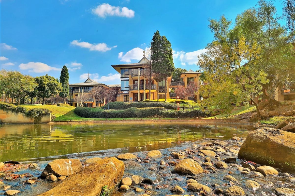 A scenic view of the office complex featuring multi-level architecture, balconies, and well-maintained landscaped gardens overlooking a tranquil pond and rocky stream.