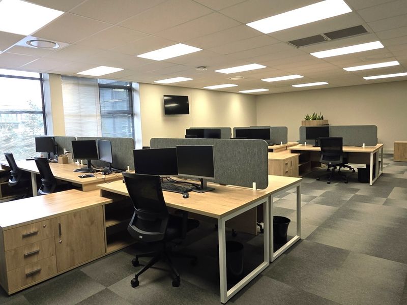 A modern, open-plan office space featuring carpet tiles, suspended ceilings with recessed lighting, and large windows providing ample natural light.