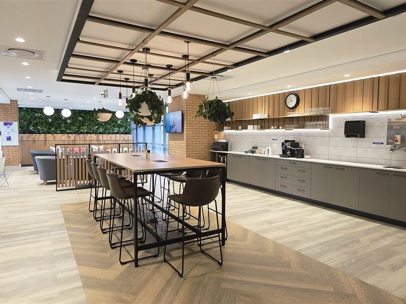 Modern open-plan office breakroom featuring a kitchenette with built-in cabinetry, a central communal table, and a stylish vertical garden wall.