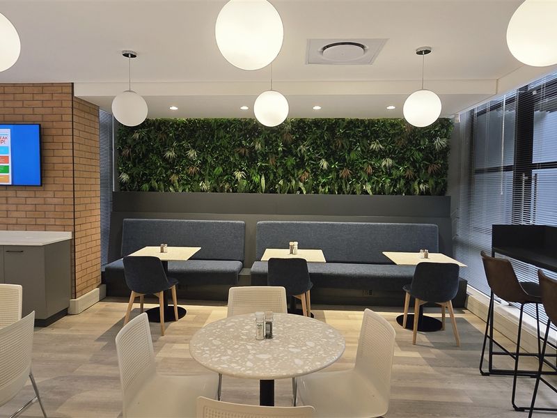Modern open-plan office breakout area featuring a vertical garden wall, built-in banquette seating, and contemporary lighting in a well-maintained professional environment.