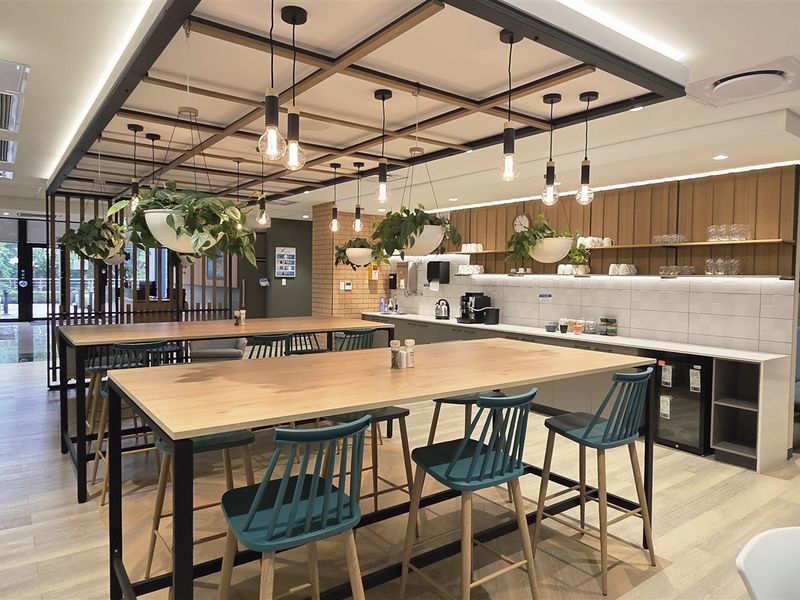 Modern open-plan office breakout area featuring a kitchenette with integrated appliances, timber-look flooring, and a stylish suspended ceiling grid with pendant lighting.