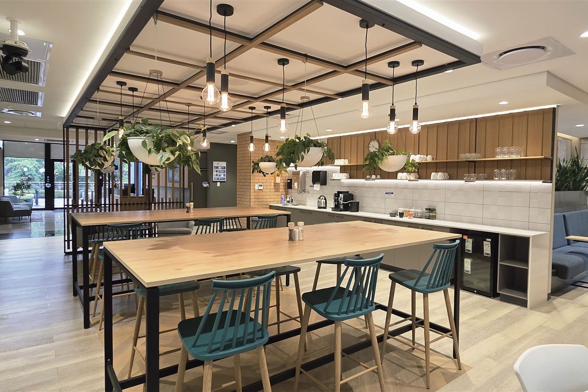 Modern open-plan office breakout area featuring a kitchenette with integrated appliances, timber-look flooring, and a stylish suspended ceiling grid with pendant lighting.
