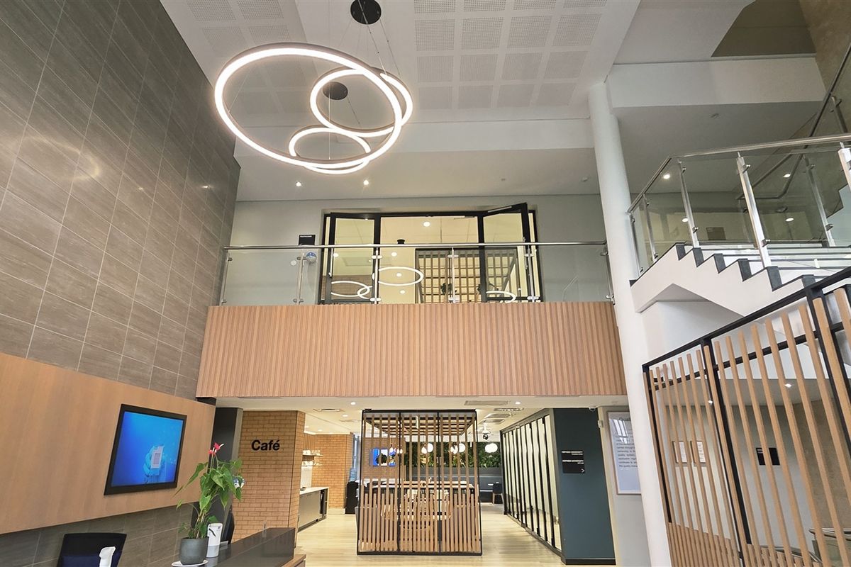 Modern double-volume office entrance lobby featuring contemporary pendant lighting, a glass balustrade staircase, and elegant timber slat wall paneling.