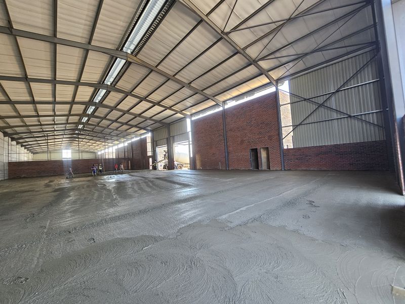 Large industrial warehouse space featuring an exposed steel portal frame, high ceilings, brick walls, and a freshly poured concrete floor.