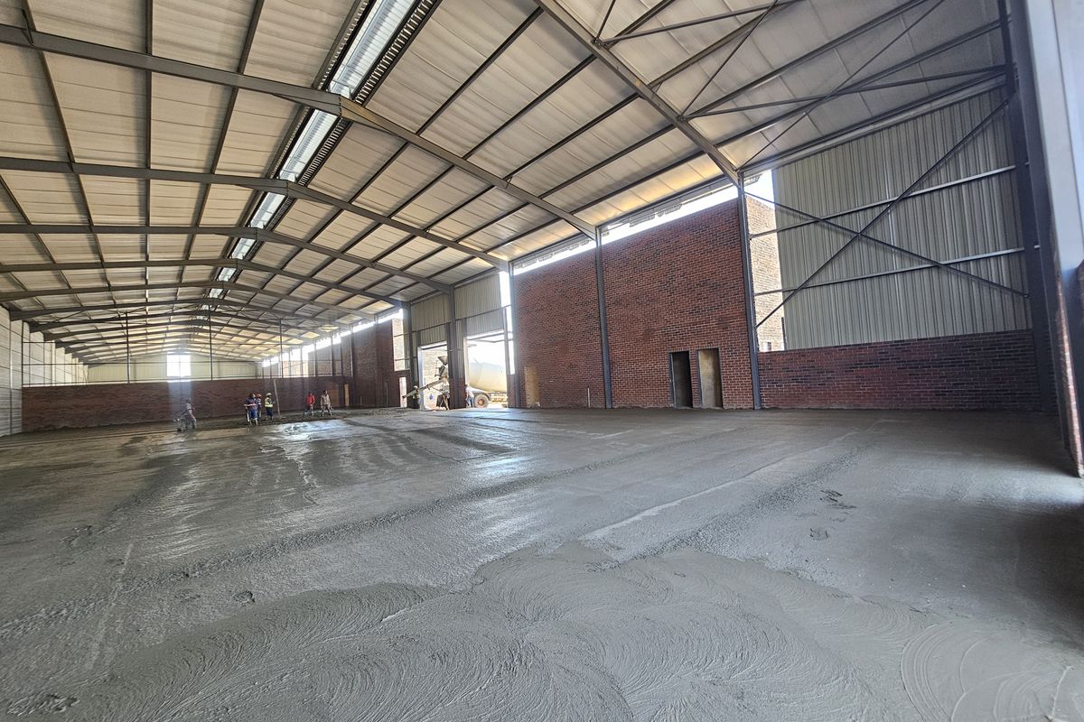 Large industrial warehouse space featuring an exposed steel portal frame, high ceilings, brick walls, and a freshly poured concrete floor.