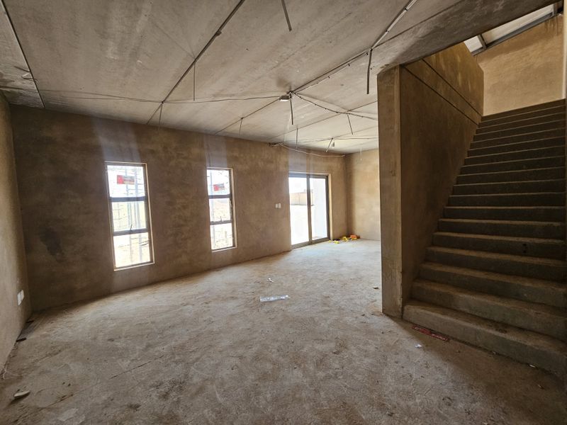 An unfinished open-plan industrial space featuring raw concrete walls, exposed ceiling, a concrete staircase, and large windows providing natural light.