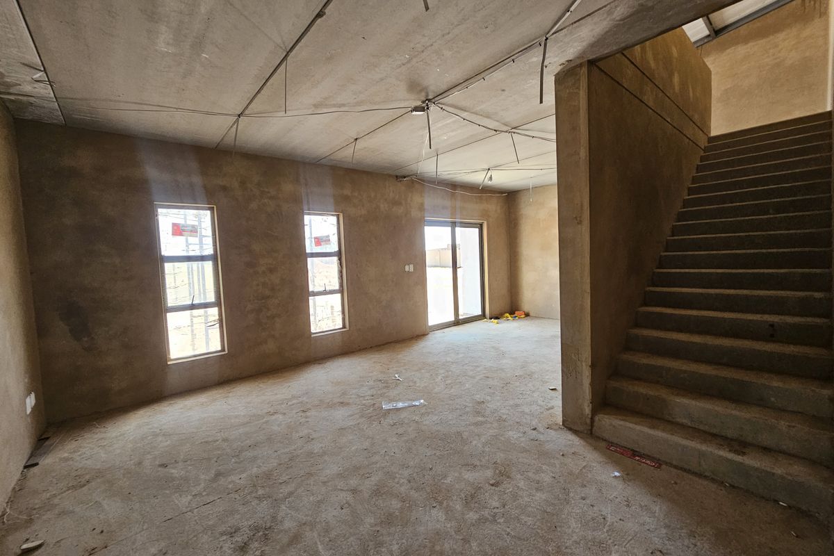 An unfinished open-plan industrial space featuring raw concrete walls, exposed ceiling, a concrete staircase, and large windows providing natural light.