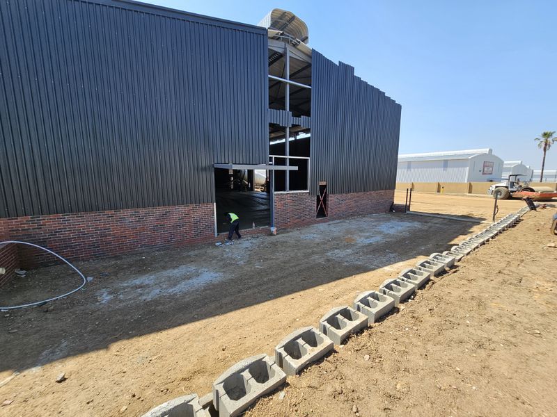 Exterior view of an industrial warehouse under construction, featuring corrugated metal cladding, brick base, and exposed steel structural framing.