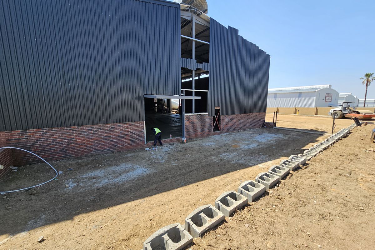 Exterior view of an industrial warehouse under construction, featuring corrugated metal cladding, brick base, and exposed steel structural framing.