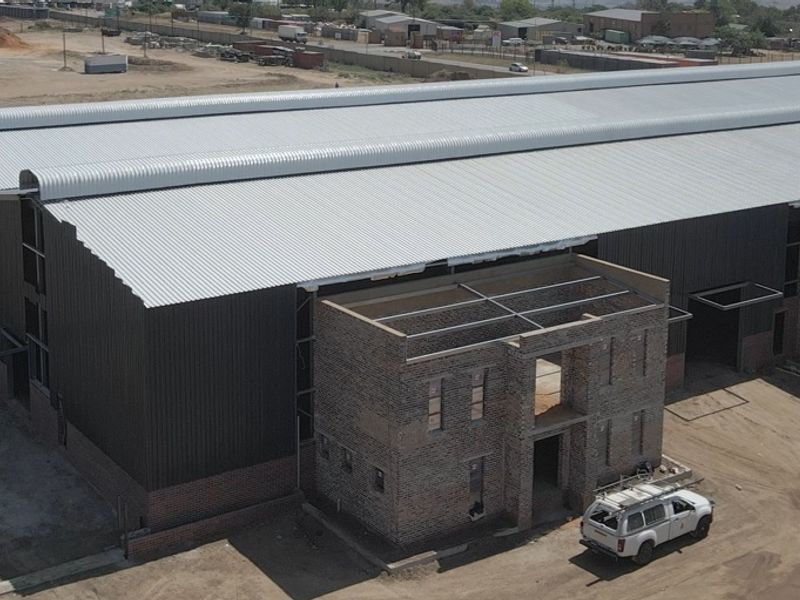 Aerial view of an industrial warehouse facility under construction, featuring a large corrugated metal roof and partially completed face brick office or utility block.