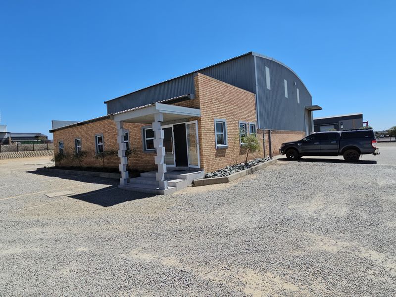 Industrial property featuring a brick facade, corrugated metal roofing, and a covered entrance, situated on a large gravel lot.