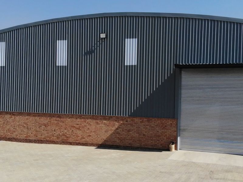 Industrial warehouse exterior featuring corrugated metal cladding, a brick base, and a large roller shutter door with paved access.