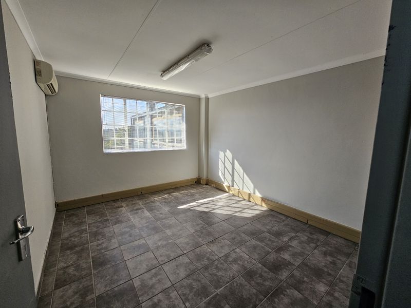 A clean, empty office space featuring tiled flooring, a wall-mounted air conditioning unit, and natural light through a window with security bars.