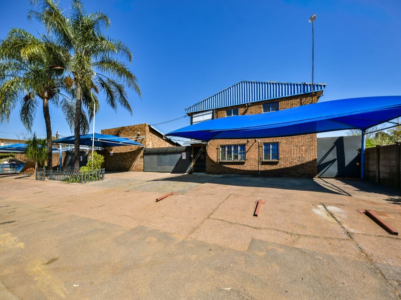 Front exterior view of an industrial property featuring a brick facade, metal roofing, and a paved parking area with shade cloth carports.