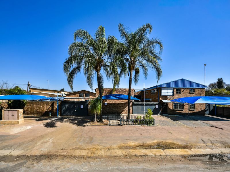 Front exterior view of an industrial property featuring a brick facade, metal roofing, paved driveway, and multiple shade cloth parking structures.