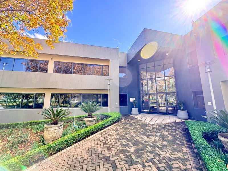 Modern multi-level office building exterior featuring a prominent glass entrance, paved walkway, and well-maintained landscaped garden areas.