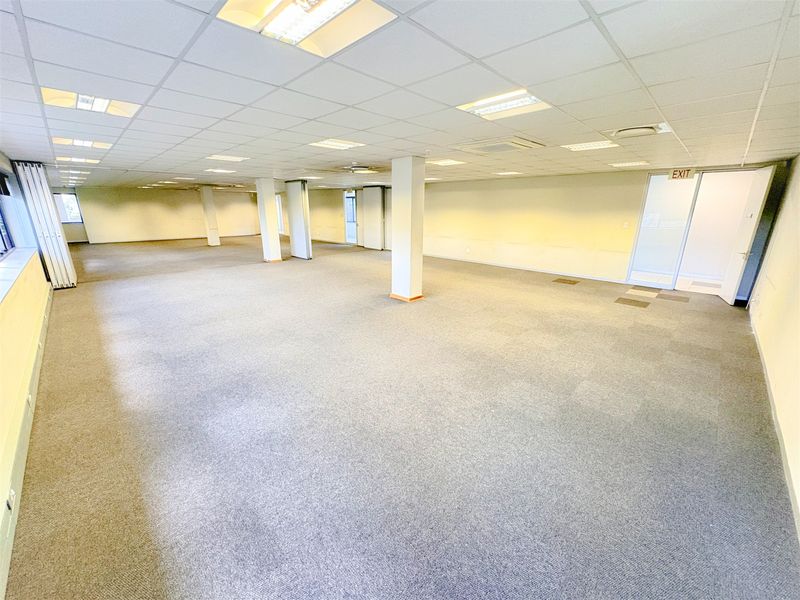 A spacious, open-plan office area featuring a suspended ceiling, recessed lighting, and neutral carpeting, offering a versatile workspace layout.