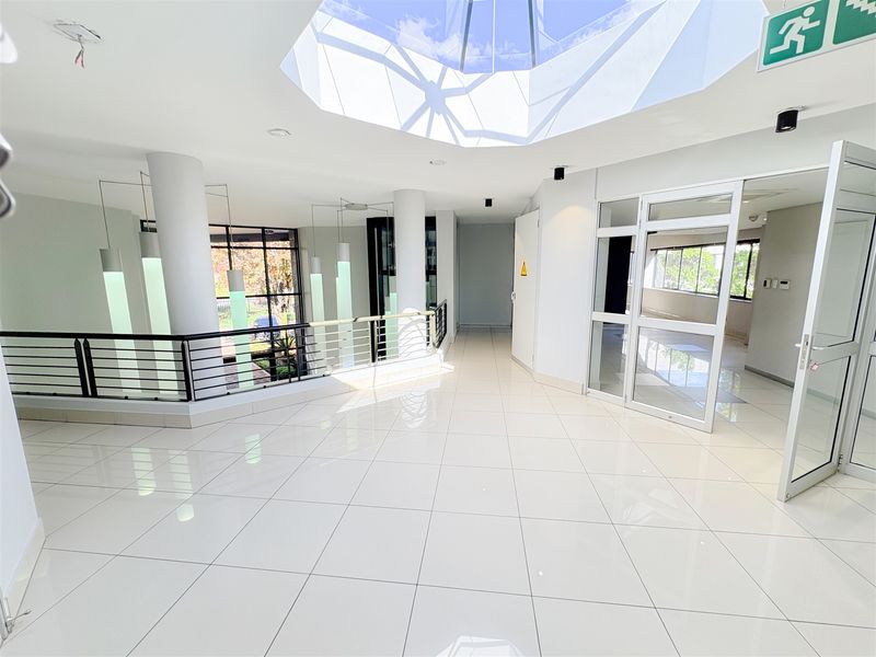 Spacious office hallway featuring polished tile flooring, a large central skylight, and glass partition doors, providing a bright and modern professional environment.