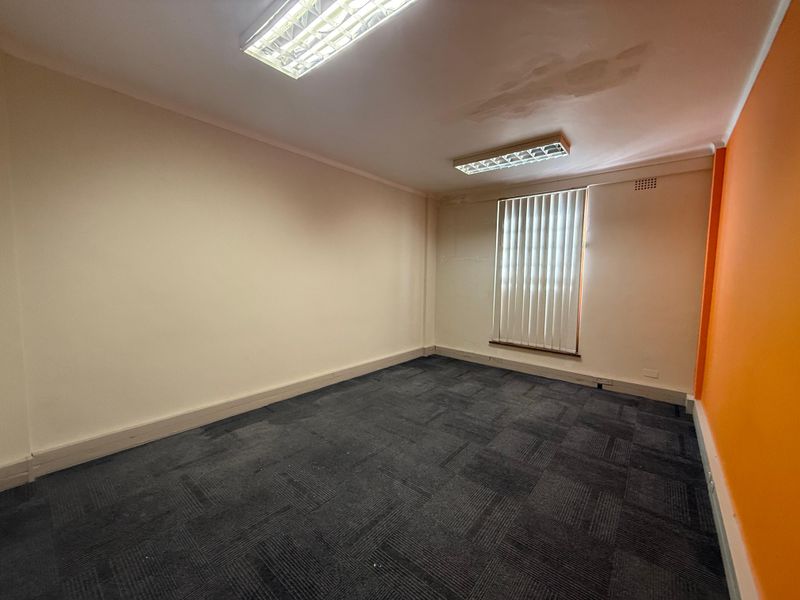 An empty commercial office space featuring dark carpet tiles, fluorescent lighting, and vertical window blinds.