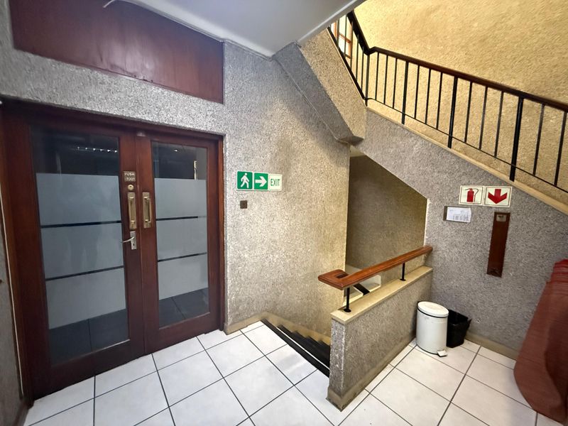 Commercial building stairwell featuring a tiled floor, metal handrails, and a secure glass-paneled wooden entrance door with emergency exit signage.