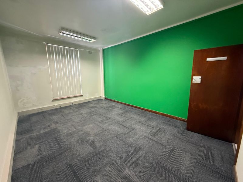 An empty commercial office space featuring carpet tile flooring, fluorescent lighting, vertical blinds, and a prominent green accent wall.
