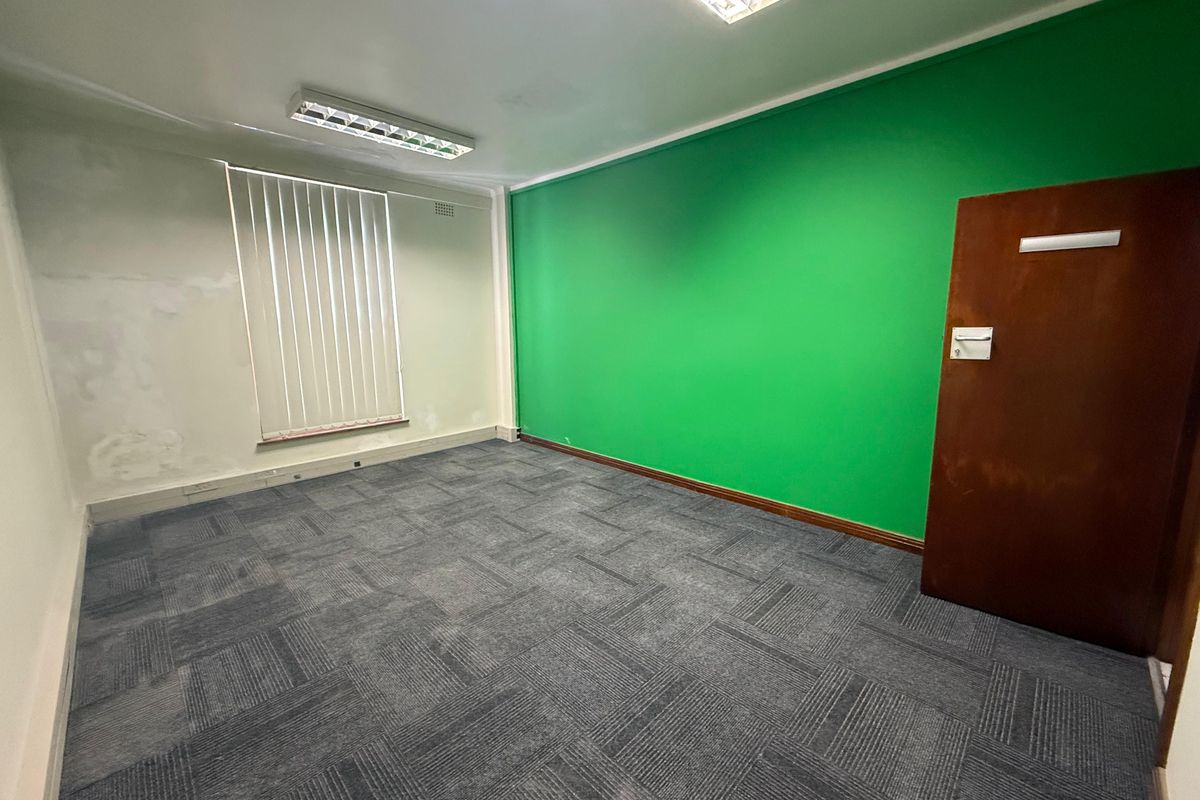 An empty commercial office space featuring carpet tile flooring, fluorescent lighting, vertical blinds, and a prominent green accent wall.