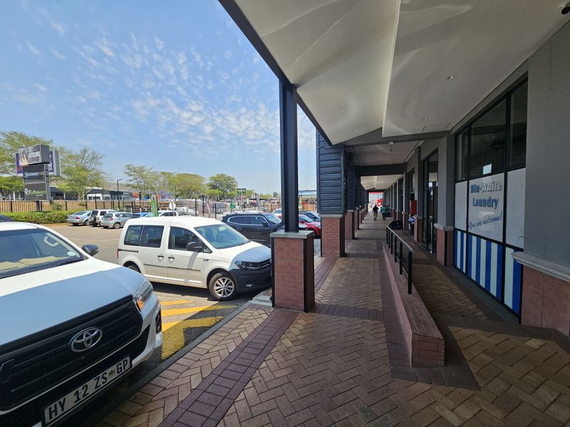 A covered walkway along the front of a retail center, featuring brick paving, structural pillars, and storefront access.