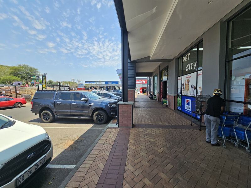 A view of the retail center walkway featuring paved flooring, covered storefronts, and convenient parking access in front of the commercial units.