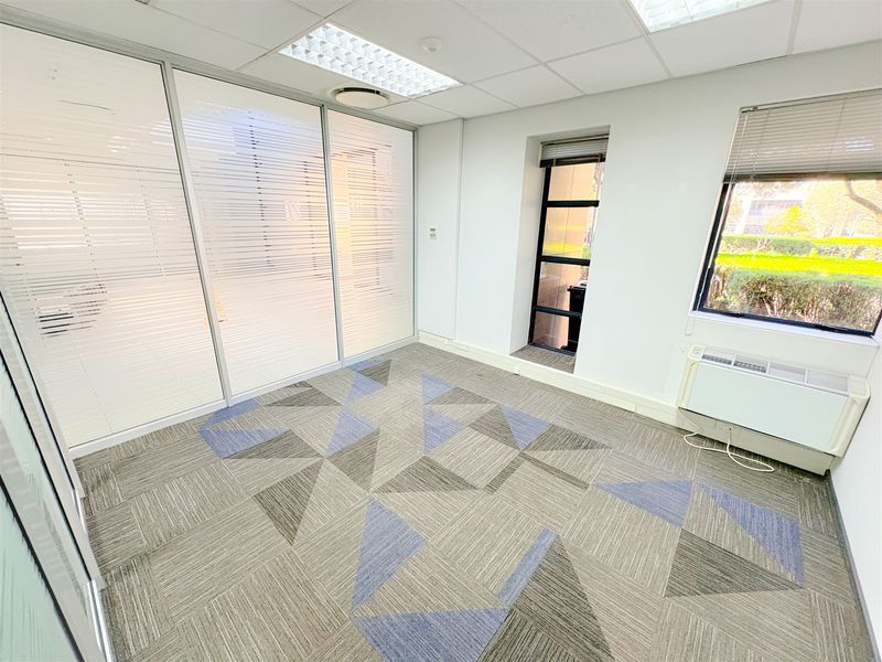 A bright, empty office space featuring modern carpet tiles, a glass partition wall, recessed ceiling lighting, and a wall-mounted air conditioning unit.