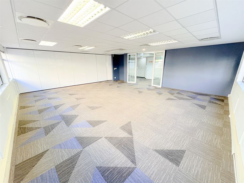 A spacious, empty office area featuring modern carpet tiles, a suspended ceiling with recessed lighting, and a large integrated whiteboard wall panel.