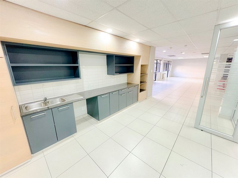 A clean, modern office kitchenette featuring built-in cabinetry, a stainless steel sink, and tiled flooring, adjacent to an open-plan workspace.