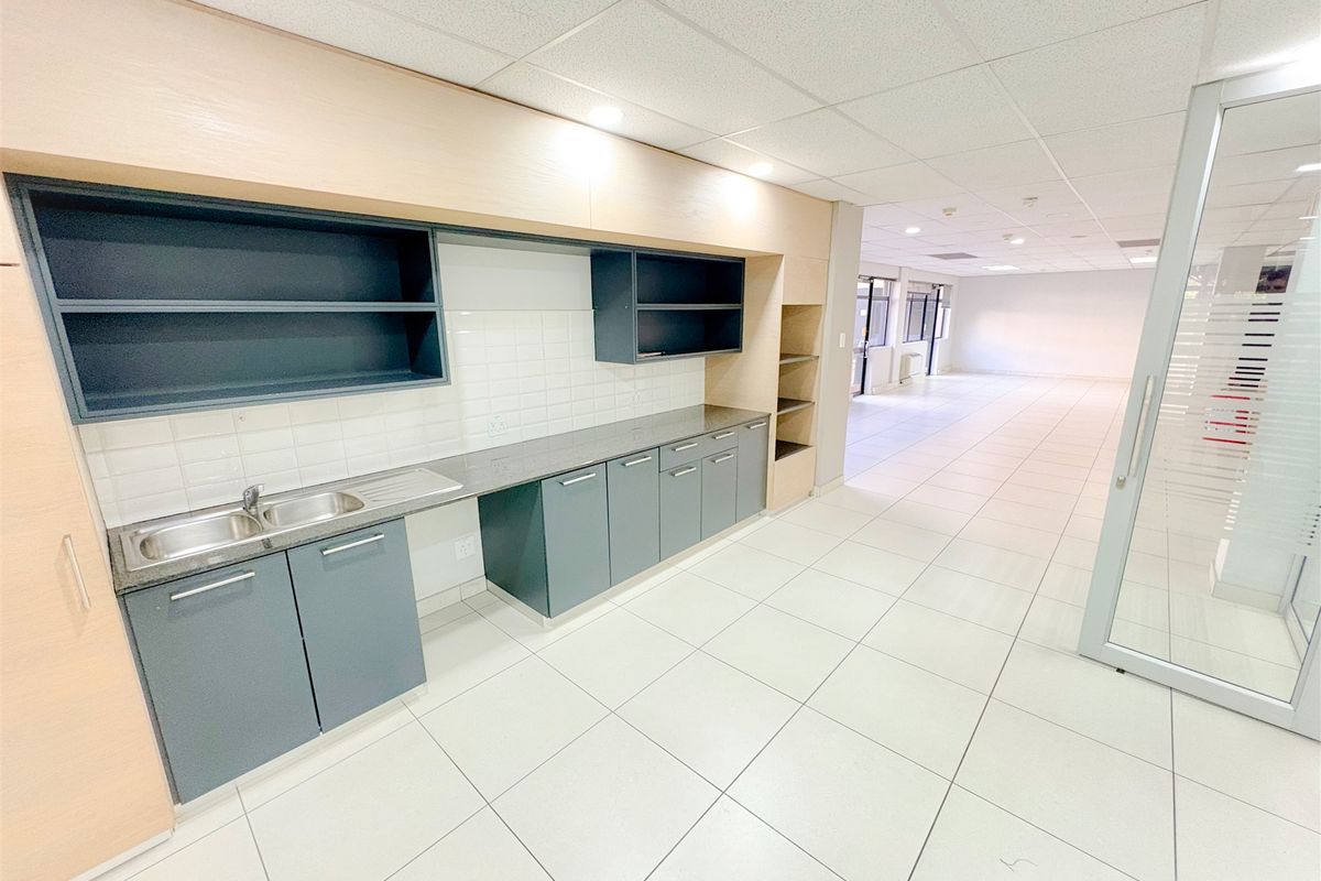 A clean, modern office kitchenette featuring built-in cabinetry, a stainless steel sink, and tiled flooring, adjacent to an open-plan workspace.