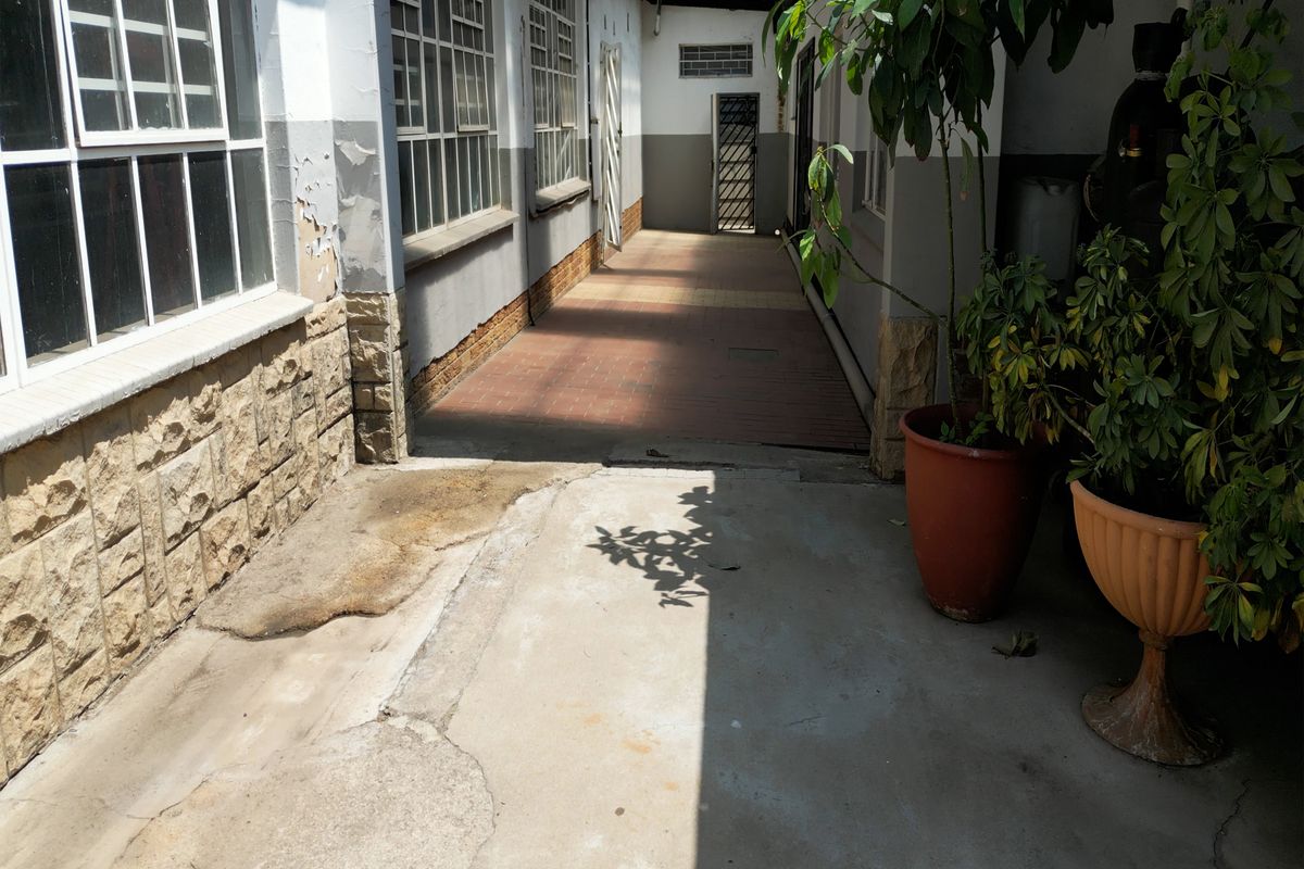 An exterior walkway area featuring concrete and tiled flooring, steel-framed windows, and masonry wall accents, showing signs of wear and weathering.