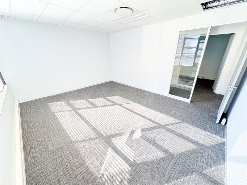 A bright, empty office space featuring modern carpet tiles, a suspended ceiling with recessed lighting, and a glass partition door.