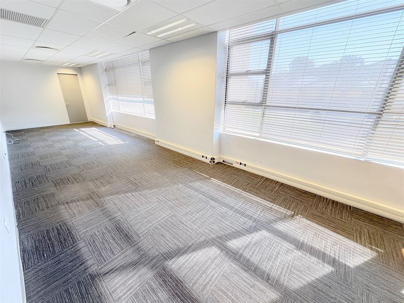 A bright, open-plan office space featuring carpet tiles, suspended ceilings with recessed lighting, and large windows with fitted venetian blinds.
