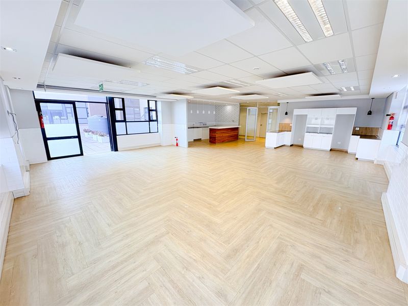 Spacious open-plan office area featuring modern herringbone flooring, suspended acoustic ceiling, and a built-in kitchenette, offering a bright and professional workspace.