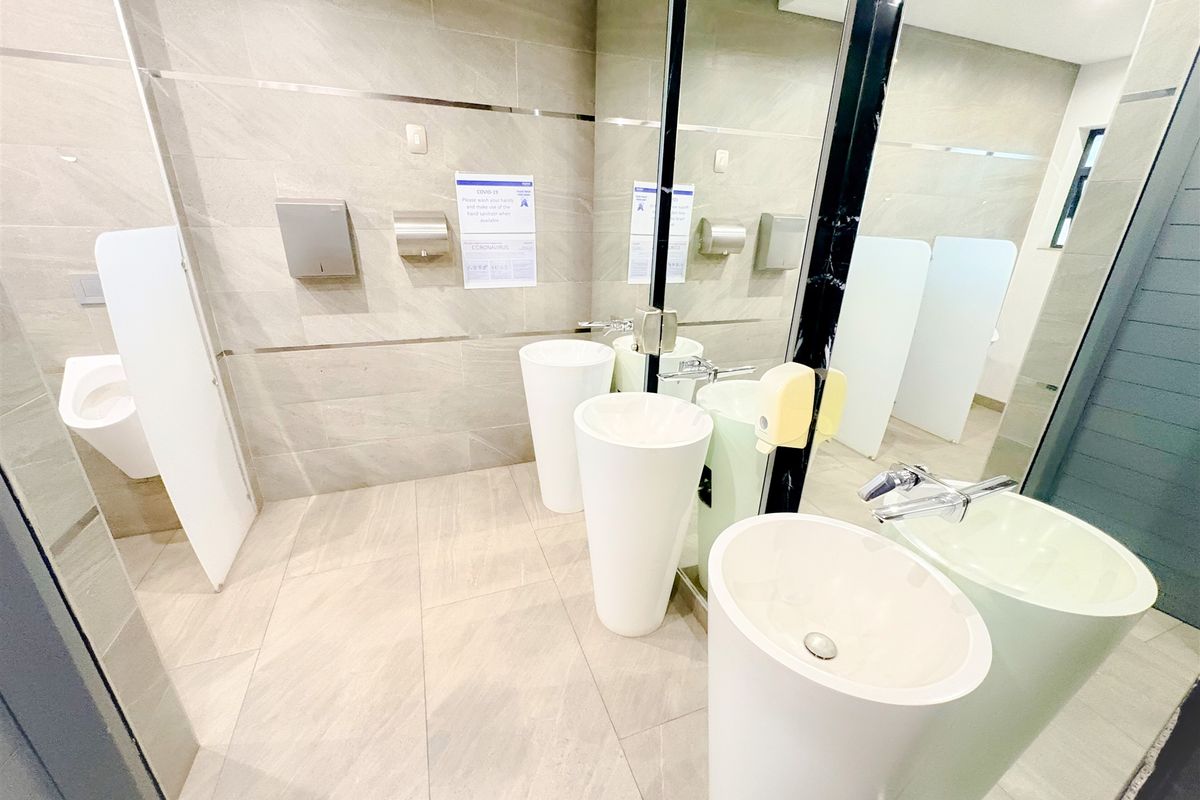 Modern office bathroom featuring pedestal washbasins, wall-mounted urinals, large vanity mirror, and neutral tiled finishes.