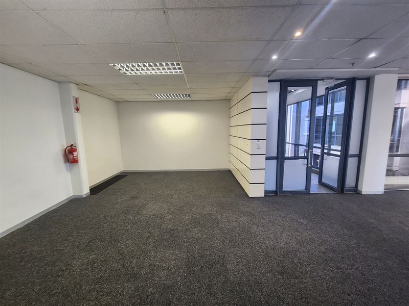 A clean, empty office space featuring a suspended ceiling, recessed lighting, and carpeted flooring with a glass entrance door.