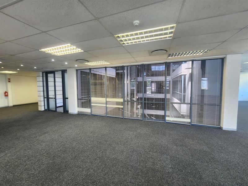 An open-plan office space featuring a suspended ceiling, recessed lighting, carpeted flooring, and a large glass partition wall overlooking an internal atrium.
