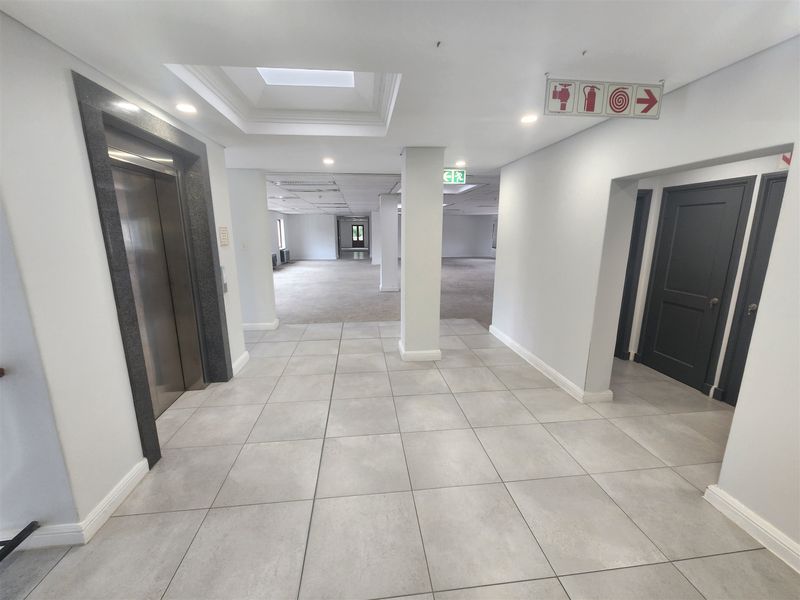A spacious, well-lit office building hallway featuring tiled flooring, a modern elevator entrance, and clear fire safety signage.